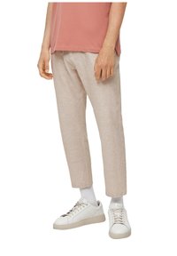 Man wearing beige linen pants, white socks, and white sneakers, standing with hands relaxed by sides.