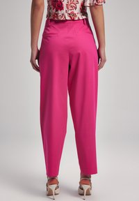 Pink tailored trousers with a tapered leg, smooth fabric, two back pockets, and a high waist. Worn with strappy high-heeled sandals.