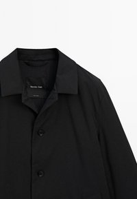 Zwarte buitenjas met een klassieke kraag, met een gladde textuur en minimalistisch design. Twee zwarte knoopsluitingen aan de voorkant.