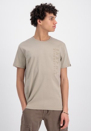 Camiseta de color beige claro hecha de algodón suave, con un sutil diseño de logo vertical en el lado izquierdo. Mangas cortas, escote redondo.