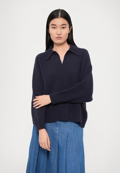 Navy blue oversized sweater with a v-neck and collar, featuring ribbed cuffs and hem. Styled with a light blue pleated skirt.