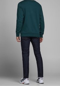 Sudadera verde con cuello redondo, combinada con jeans oscuros. El calzado tiene una base blanca con acentos azules y naranjas.