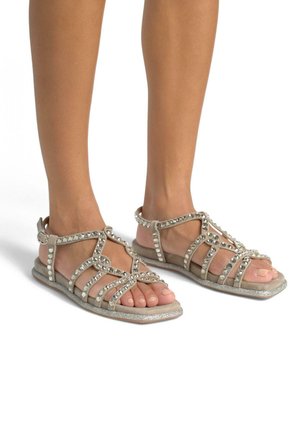 Sandali beige con più cinturini abbelliti da rhinestone argentati. Suola piatta con leggera lucentezza e cinturino alla caviglia per supporto. Design open-toe.