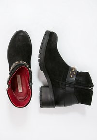 Black suede ankle boots with a chunky black heel, featuring gold stud accents and a side zip closure. Red interior lining.