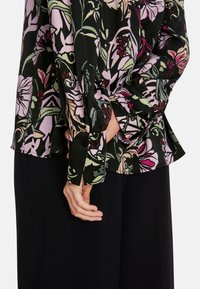 Bluse mit floralen Mustern in Schwarz mit grünen, rosa und cremefarbenen Akzenten. Hat lange Ärmel und knöpfbare Manschetten. Aus weichem, leichtem Material.