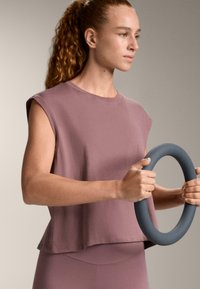 Bluza lejeră cu mânecă scurtă dintr-un material mauve estompat, combinată cu pantaloni mulați asortați. Modelul ține un inel de exerciții gri.