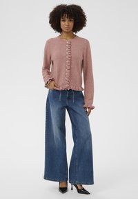 Világos rózsaszín kardigán fodros előlappal és gombos záródással. Széles lábú kék farmer nadrággal és fekete hegyes orrú magassarkúval párosítva.