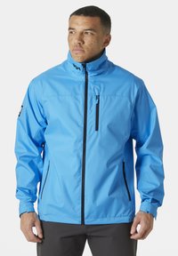 Chaqueta impermeable azul con un alto cuello, cierre frontal con cremallera y dos bolsillos laterales con cremallera. Cuenta con una textura suave y puños ajustables.