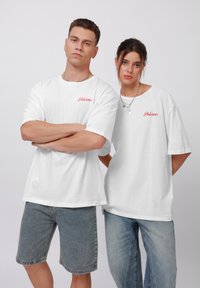 Hvide kortærmede T-shirts med rød "Palermo" tekst. En person har denimshorts på; den anden har lyseblå jeans. Enkel design, afslappet pasform.