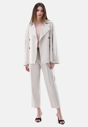 Junge Frau trägt beige zweireihigen Blazer, passende hoch taillierte Hose, hellrosa Top und schwarze offene Zehenabsätze, steht vor einfachem Hintergrund.