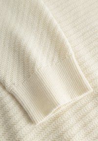 Ecrufarbenes Strickgewebe mit Fischgrätenmuster, mit geripptem Bündchen, das eine weiche Textur zeigt.