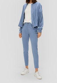 Mujer vestida con una chaqueta con cremallera azul claro, camisa blanca, pantalones azules a juego y zapatillas blancas, de pie contra un fondo claro liso.