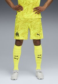 Il completo da calcio giallo brillante presenta un design testurizzato, loghi neri sui pantaloni e sulle calze, e scarpini bianchi. Le calze sono alte fino al ginocchio con un motivo a righe.