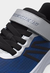 Μπλε και μαύρο αθλητικό παπούτσι με μαύρα κορδόνια και ασημί λουράκι με βέλκρο με την ένδειξη "Skechers" πάνω από τη γλώσσα.