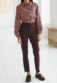 Chemise à boutons rose avec un motif abstrait noir ; pantalon taille haute bordeaux foncé ; mocassins noirs brillants. Tissu lisse et coupe ajustée.