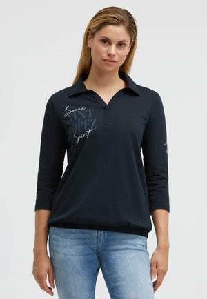 Woman wearing a navy blue long-sleeve polo shirt with text "Soccx Saint Tropez Spirit" paired with light blue jeans, standing against a white background.