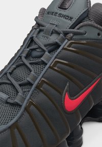 Temno siva športna copatka Nike Shox z rdečim logotipom, mrežastim zgornjim delom, elastičnim vezanjem in vidno oblazinjeno podplatom.