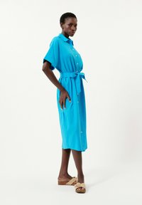Blaues Hemdkleid mit kurzen Ärmeln, tailliertem Design, knielanger Länge und glatter Textur. Getragen mit hellen Plateausandalen.
