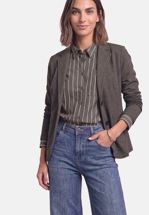 Donna che indossa un blazer scuro sopra una camicia a righe con bottoni e jeans blu, una mano in tasca, in piedi contro uno sfondo chiaro e uniforme.