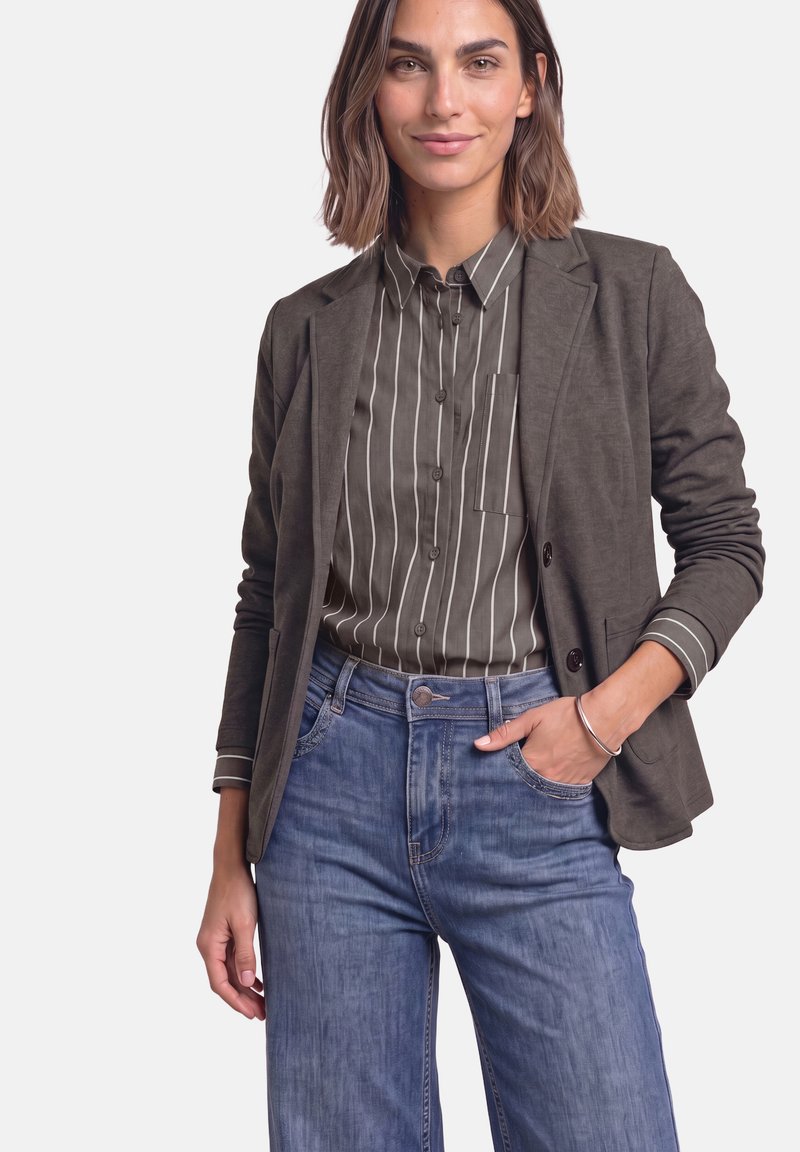 Femme portant un blazer foncé par-dessus une chemise rayée avec boutons et un jean bleu, une main dans la poche, debout devant un fond clair uni.