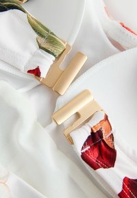 Valge kangas lillemotiividega, millel on kuldsed riistvara kinnitusdetailid, sile tekstuur ja rihmal voltidega osa.