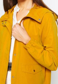 Żółta lekka kurtka z kapturem, z przodem zapinanym na zamek błyskawiczny, srebrnymi zatrzaskami, elastycznymi mankietami i boczną kieszenią. Gładka faktura.