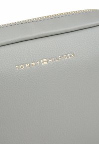 Borsa in pelle grigia con texture liscia e logo oro "TOMMY HILFIGER". Dettaglio ravvicinato della cerniera lungo il bordo superiore.