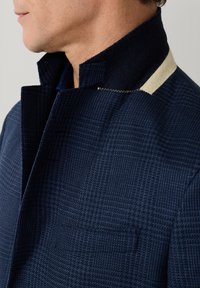 Mann trägt einen marineblauen karierten Mantel mit hochgestelltem Kragen und einem beigen Stoffdetail auf der Schulter.