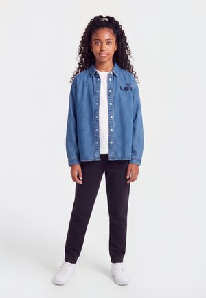Fille aux longs cheveux bouclés attachés, portant une veste en jean bleue, une chemise blanche, un pantalon noir et des baskets blanches, debout sur un fond uni.