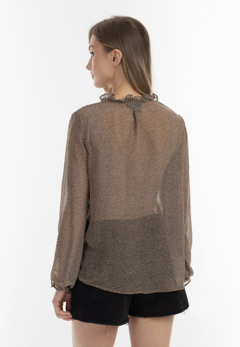 Junge Frau mit braunem Haar, die eine durchsichtige, langärmlige, braun gemusterte Bluse und schwarze Denim-Shorts trägt, mit dem Rücken zu einer weißen Hintergrund.