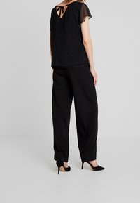 Black top with a textured, glittery finish and short, sheer sleeves; features a tied back neckline. Paired with loose black trousers and heels.