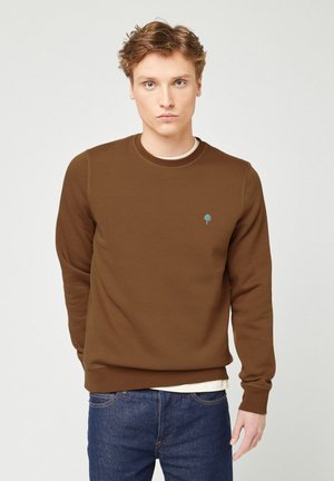 Giovane uomo con capelli castano chiaro che indossa un felpa marrone con un piccolo emblema di un albero blu, sopra una maglietta bianca e jeans blu, in piedi.