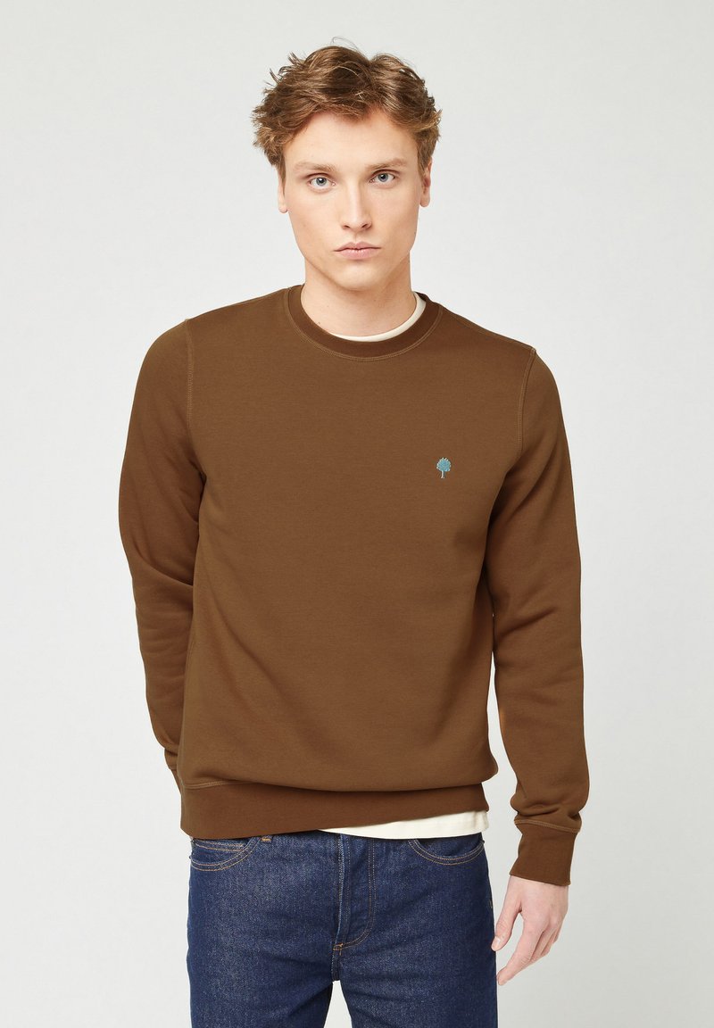 Giovane uomo con capelli castano chiaro che indossa un felpa marrone con un piccolo emblema di un albero blu, sopra una maglietta bianca e jeans blu, in piedi.
