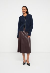 Moteris vilkinti tamsiai mėlyną užtraukiamą pūkuotą kardiganą, juodą viršutinį drabužį, tamsiai rudą odinę midi sijoną ir juodas žemas kulniuko batus prieš baltą fono.