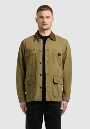 Mężczyzna w beżowej kurtce zapinanej na guziki z czterema przednimi kieszeniami i ciemnym kołnierzem, noszący czarną koszulę i czarne spodnie, stojący na jednolitym tle.