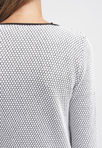 Biały sweter z teksturowanym wzorem małych czarnych heksagonów, z zaokrąglonym dekoltem i kontrastową, czarną lamówką wzdłuż dekoltu.