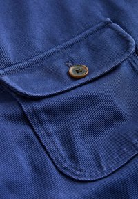 Marineblåt stof med ribbet tekstur, der har en klaplomme med en rund brun knap i midten. Rene syningsdetaljer er synlige.