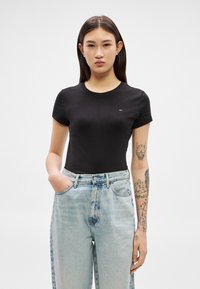 Ung kvinde med langt mørkt hår, iført en tætsiddende sort t-shirt og lyseblå højtaljede jeans, stående med den ene hånd i lommerne.