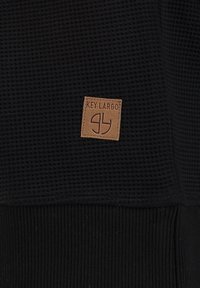 Maglione nero con texture, orlo e polsini a coste; presenta un'etichetta quadrata color marrone con la scritta "KEY LARGO" e un logo nell'angolo in basso.