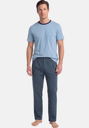 Uomo in piedi a piedi nudi che indossa una camicia azzurra a maniche corte con tasca e pantaloni da pigiama blu scuro a fantasia su sfondo semplice.
