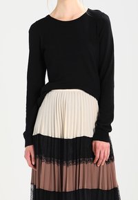 Svart långärmad tröja i kombination med en veckad kjol som har horisontella ränder i grädde, brunt och svart, accentuerad med spets.