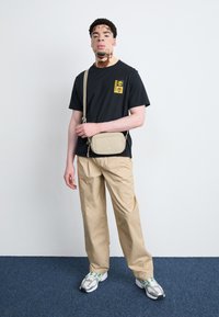 Zwarte t-shirt met gele logo, beige schoudertas, beige broek en grijze sneakers met groene accenten. Eenvoudige achtergrond en blauwe tapijt.