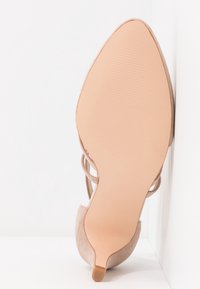 Chaussure beige à talon haut à bout pointu montrée de la semelle inférieure avec un talon fin et des brides fines sur fond blanc.