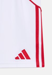 Ropa deportiva blanca con rayas rojas y un logo de Adidas bordado en rojo en un diseño triangular en el dobladillo. Textura de tela suave.