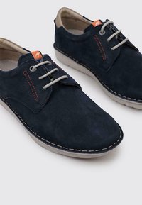 Zapatos de ante azul marino con cordones grises, detalles de costura y una suela beige. Presentan un corte lateral y una pequeña etiqueta naranja cerca de los cordones.