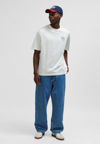 White t-shirt with a round neck and short sleeves, paired with loose blue jeans. A navy and red cap completes the outfit. White sneakers visible.