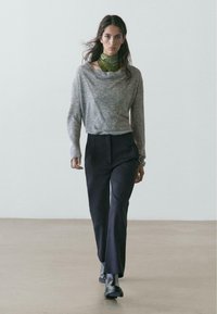 Maglione grigio chiaro con scollo drappeggiato, abbinato a pantaloni scuri e stivaletti neri. Sciarpa verde con motivo intorno al collo che aggiunge colore.
