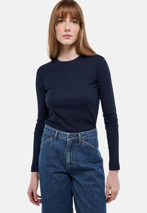 Femme aux cheveux brun clair et raides, portant un haut à manches longues bleu marine et un jean taille haute bleu, debout devant un fond blanc uni.