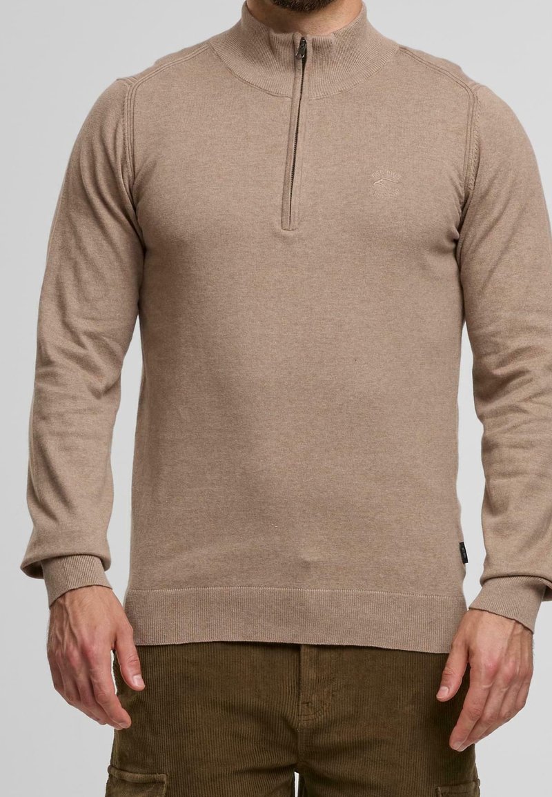 Homme portant un pull en maille beige à manches longues avec col demi-fermeture éclair et pantalon en velours côtelé marron, montré du milieu de la poitrine jusqu’aux hanches.