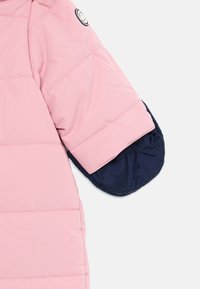 Rosa quiltet jakkeerm med marineblåt inderside og en sirkulær svart-hvitt logo-patch nær skulderen.